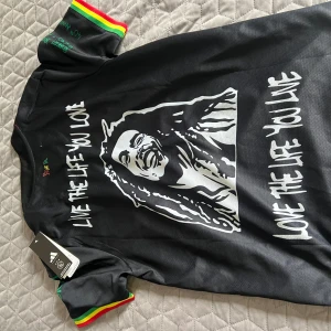 Ajax x Bob Marley svart fotbollströja - Unik svart fotbollströja från Ajax och Adidas med Bob Marley-tema. Tröjan har coola tryck med Bob Marleys ansikte, citat och färgglada detaljer i rött, gult och grönt på ärmarna. Kortärmad modell i lätt och ventilerande material, perfekt för match eller streetwear.