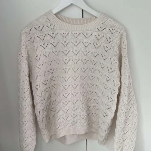 Beige stickad tröja med hålmönster - Säljer en beige stickad tröja med snyggt hålmönster i geometriska former. Tröjan har rund hals och långa ärmar med ribbade muddar. Perfekt för dig som gillar en chill och stilren look med lite extra detaljer.