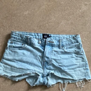 Ljusblå jeansshorts från 157 - Snygga ljusblå jeansshorts från 157 med låg midja och råa, fransiga kanter. Klassisk femficksmodell med dragkedja och knapp framtill. Perfekta för sommardagar och ger en avslappnad vibe.
