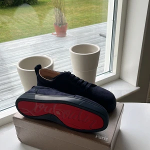Mörkblå sneakers från Christian Louboutin - Snygga mörkblå sneakers från Christian Louboutin med lyxig mocka och klassisk röd sula. Skorna har rund tå, platt sula och snörning framtill. Perfekt för dig som vill sticka ut med exklusiv stil och detaljer som verkligen syns. Helt oanvända.