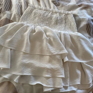 Vit volangkjol med smock - Supersöt vit kjol med bred smock i midjan och dubbla volanger som ger en flowig och lekfull look. Kjolen är kort och har en mjuk, lätt struktur i tyget som känns skönt mot huden. Perfekt för dig som gillar romantiska vibes och vill sticka ut.