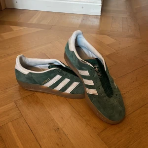 Adidas Gazelle gröna sneakers strl 40⅔ - Gröna Adidas Gazelle sneakers med klassiska vita ränder och gummisula. Ovandel i mocka och vit insida, med guldfärgad Gazelle-text på sidan. Snygg retrostil och låg profil, perfekt för dig som gillar streetwear och ikoniska modeller.