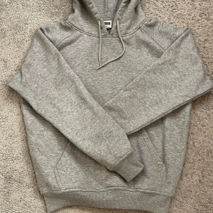 Grå hoodie från Urban Classics XS - Säljer en stilren grå hoodie från Urban Classics i storlek XS. Tröjan har huva med snörning, ribbade muddar och en klassisk känguruficka framtill. Perfekt för chill dagar och enkel att matcha med jeans eller joggers. Oandvänd så skicket är 10/10
