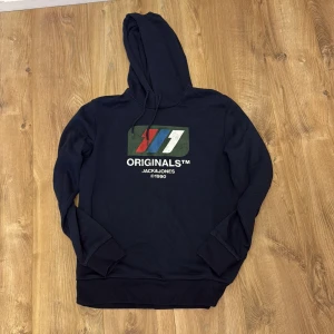 Mörkblå hoodie Jack & Jones Originals - Snygg mörkblå hoodie från Jack & Jones Originals med tryck i rött, blått, vitt och grönt framtill. Tröjan har huva med snörning och långa ärmar. Perfekt för chill dagar och streetstyle. Materialet känns som mjuk bomull och passformen är normal.