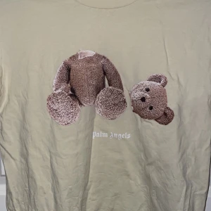 Beige t-shirt med nalletryck - t-shirt i beige med stort tryck av en dekonstruerad nalle på framsidan och Palm Angels-logga i vitt på både fram- och baksidan. Klassisk rund hals och korta ärmar. Materialet är mjuk bomull, perfekt för en chill streetwear-look.