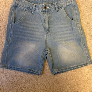 Ljusblå jeansshorts från Carhartt - Säljer ett par ljusblå jeansshorts från Carhartt med klassisk femficksdesign och bälteshällor. Shortsen har raka ben, knapp och dragkedja framtill samt Carhartt-logga på ena bakfickan. Perfekt för chill dagar och enkel att matcha med t-shirt eller hoodie. Passar som strolek (S/M)