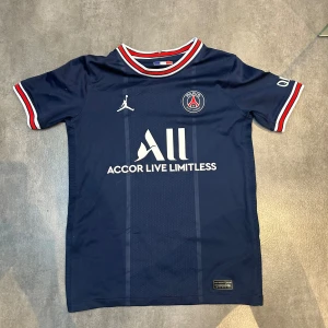 PSG matchtröja marinblå Jordan - Snygg PSG matchtröja i marinblått med vita och röda detaljer på krage och ärmslut. Tröjan har korta ärmar, PSG-logga på bröstet och Jordan-logga. Materialet är lätt och andas, perfekt för fotboll eller häng. Sponsorn Accor Live Limitless tryckt på framsidan.