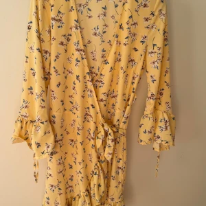 Gul blommig omlottklänning med volang - Superfin gul omlottklänning med blommigt mönster och volanger. Klänningen har långa ärmar med knyt och v-ringad hals. Sitter snyggt med knyt i midjan och har en flowy