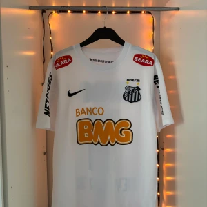 Santos Neymar Jr 11 hemmatröja Nike - Säljer en  Santos FC fotbollströja med Neymar Jr och nummer 11 på ryggen. Tröjan är vit med svarta och orangea tryck, har korta ärmar och är tillverkad i lätt Dri-Fit material. Flera sponsorer och klubbmärke på bröstet. Perfekt för dig som älskar brasiliansk fotboll.