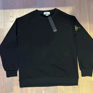 Svart sweatshirt från Stone Island - Svart sweatshirt från Stone Island med klassisk rund hals och ribbade muddar. Ikonisk logotyp-patch på vänster ärm i gult och grönt. Tröjan har en clean och stilren look, perfekt för dig som gillar streetwear. Materialet är mjuk bomull för skön komfort.
