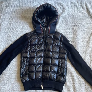 Svart Moncler cardigan med huva - Svart cardigan från Moncler med quiltad framsida och huva. Ärmarna och ryggen är stickade, vilket ger en schysst kontrast mot det blanka materialet på kroppen. Jackan har dragkedja framtill och Moncler-logga på ena ärmen. Perfekt för dig som gillar stilren design. 