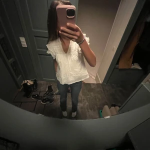 Vit blus med volang från Gina Tricot - Supersöt vit blus från Gina Tricot med v-ringning och korta ärmar med volangdetaljer. Blusen har en luftig passform och fina broderade detaljer upptill. Perfekt för dig som gillar en romantisk och chill stil.