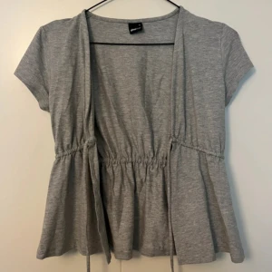 Grå omlottopp med knyt från Gina Tricot - Säljer en ljusgrå omlottopp från Gina Tricot med korta ärmar och knyt i midjan. 