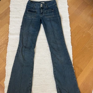 Blå bootcut jeans från Nelly - Snygga blå jeans från Nelly med bootcut passform och låg midja. Jeansen har två framfickor , dubbla knappar i midjan och markerade sömmar längs benen. Materialet är klassisk jeans med lite stretch för skön passform.