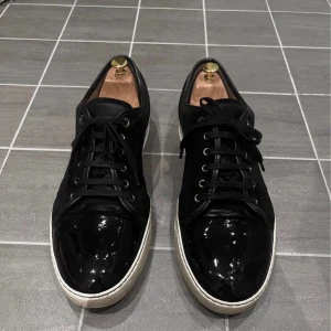 Lanvin skor - Säljer ett par svarta Lanvin sneakers med snygg lackad tå och vit platt sula. Skorna har snörning och en stilren design med rund tå. Materialet är en mix av mocka och lack, vilket ger en lyxig känsla. Perfekt för dig som gillar exklusiva och trendiga sneakers, storlek 43