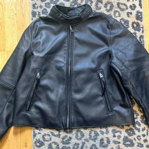 Snygg jacka från H&M - En helt ny svart bikerjacka med silverfärgade detaljer i ytbehandlad jättefin kvalitet. Jackan har ståkrage med tryckknappar, dold dragkedja fram och sidfickor med dragkedja. Lång ärm med dragkedja vid ärmslut. Den är fodrad med ett glansigt innerfoder Jackan är lite utställd i modellen Storlek large men passar storlek 36-40 Längd: 62cm Bredd under ärm: 61x2cm Ärmlängd: 63cm Nypris 699:-