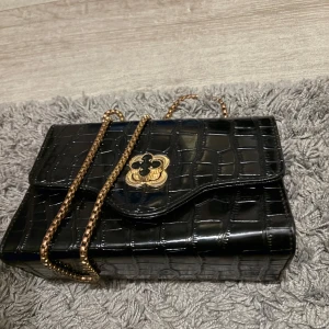 Svart axelväska med kedja  - Snygg svart axelväska med krokopräglad yta och guldfärgad kedja. Väskan har ett dekorativt blomformat spänne i guld och svart, samt ett praktiskt innerfack med dragkedja. Perfekt för att lyfta din outfit med lite extra edge.