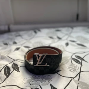 Louis Vuitton bälte  - Snyggt svart skärp från Louis Vuitton med klassiskt monogrammönster och stor LV-logga i silver på spännet. Bältet är gjort i skinn och har en brun insida. Perfekt accessoar för att lyfta din outfit med lyxig känsla.