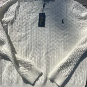 Vit kabelstickad tröja Polo Ralph Lauren - Vit kabelstickad tröja från Polo Ralph Lauren med klassisk rund hals och broderad marinblå logga på bröstet. Tröjan har långa ärmar och ribbade muddar. Perfekt för dig som gillar stilren och tidlös design. Står att den är L me  sitter som M, långa armar, kort midja.