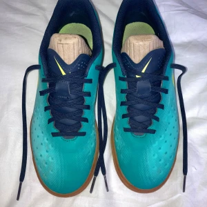 Nike blå fotbollsskor med gummisula - Säljer ett par Nike fotbollsskor i blått med stora gula Swoosh-loggor på sidorna. Skorna har marinblå snörning och detaljer, samt en platt, brun gummisula med mönster för bra grepp inomhus. Ovandelen är i syntetmaterial med små prickar för extra stil.