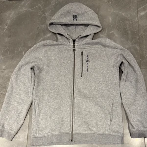 Grå hoodie från Sail Racing med dragkedja - Snygg grå hoodie från Sail Racing med hel dragkedja och huva med snörning. Tröjan har en bröstficka med dragkedja och logga på vänster sida. Insidan är fodrad med mjuk fleece för extra komfort. Perfekt för chill dagar eller när du vill vara extra bekväm.