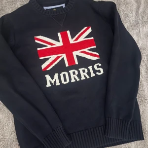Morris stickad tröja med flagga - Snygg mörkblå stickad tröja från Morris i storlek medium. Stor Union Jack-flagga i rött, vitt och blått på bröstet samt Morris-logga under. Tröjan har rund hals och ribbade muddar vid ärmslut och nederkant. Perfekt för dig som gillar klassisk stil med en twist.