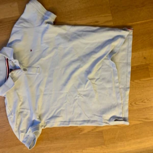 Ljusblå piké från Tommy Hilfiger - Fräsch ljusblå pikétröja från Tommy Hilfiger med klassisk krage och två knappar framtill. Diskret logga broderad på bröstet och snygga ränder i rött, vitt och blått på insidan av kragen. Perfekt för en clean och stilren look.