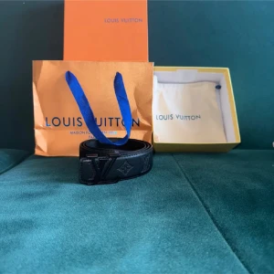 Svart loi s vitten skärp i skinn - Säljer ett svart  bälte i äkta skinn med ikonisk L. V-logga på spännet och präglade monogramdetaljer längs hela bältet. Stilren och lyxig design som lyfter din outfit. Kommer med originalförpackning och dustbag.