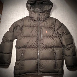 Mörkgrön pufferjacka Polo Ralph Lauren - Säljer en mörkgrön  pufferjacka från Polo Ralph Lauren med huva och orange broderad logga på bröstet. Jackan har flera dragkedjor, quiltad design och långa ärmar. Använd i en säsong och inga slitage siter som xs-s fast