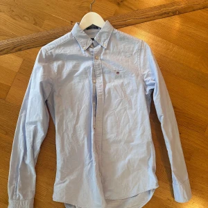 Ljusblå skjorta från GANT - Snygg ljusblå skjorta från GANT med klassisk krage och knappar framtill. Skjortan har lång ärm och en liten broderad logga på bröstet. Perfekt för dig som gillar stilrena och tidlösa plagg. Materialet är mjuk bomull som känns skönt mot huden.