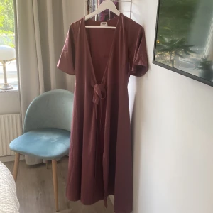 Vinröd långklänning från Bubbleroom - Supersnygg vinröd långklänning från Bubbleroom med korta ärmar och v-ringad omlottfront. Klänningen har ett knyt i midjan som ger en fin siluett och är tillverkad i ett mjukt, lätt material. Perfekt för dig som gillar stilrena och feminina plagg.