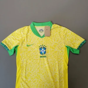 Brasilien retreo Nike gul fotbollströja - Snygg Brasilien fotbollströja från Nike i gul med gröna detaljer och grafiskt mönster. Kortärmad med V-ringad grön krage och blå/gröna ränder på ärmsluten. CBF-logga och Nike Swoosh på bröstet. Perfekt för match eller häng med kompisarna.