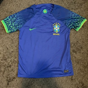 Blå Brasil Nike fotbollströja - Säljer en blå Brasilien fotbollströja från Nike med coolt leopardmönster i grönt på ärmarna och gula detaljer vid mudden. Tröjan har Brasil-logga med fem stjärnor på bröstet och är tillverkad i lätt, ventilerande polyester. Perfekt för dig som älskar fotboll och snygg design.