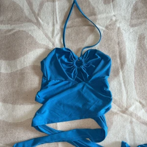 Blå omlott halterneck topp - Snygg blå omlott topp med halterneck och knytband som går runt midjan. Toppen har en cool rynkad detalj framtill och är tillverkad i mjukt, stretchigt material. Perfekt för dig som vill ha en trendig och unik look med öppen rygg.