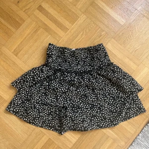 Svart blommig volangkjol - Supersöt svart kortkjol med vita små blommor och volanger. Kjolen har en smockad midja som ger en skön och flexibel passform. Har shorts under. Skriv vid minsta fråga!💕💕är storlek 152 men skulle passa en xxs-xs