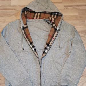 Grå Burberry hoddie  - Grå hoodien från Burberry med klassiskt rutigt foder i huvan och broderad logga på bröstet. Jackan har dragkedja, snörning i huvan och två fickor framtill. Perfekt för dig som gillar stilren design med en touch av lyx.