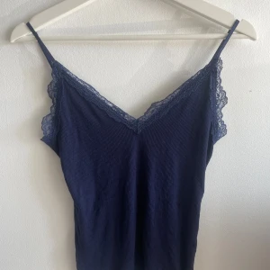 Mörkblå linne med spets från new yorker - Supersnygg mörkblå linnetopp från new yorker med tunna axelband och v-ringad hals. Toppen har fina spetsdetaljer längs kanten och är ribbad i materialet. Använd få gånger och har inget slitage