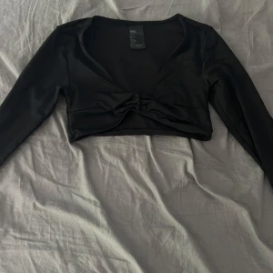 Svart croppad topp från H&M - Svart långärmad croppad topp från H&M med twistad detalj framtill. Tunn och stretchig polyester som sitter snyggt och markerar midjan. Perfekt för dig som gillar trendiga och stilrena plagg med en cool touch.