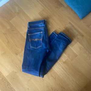 Blå jeans från Nudie Jeans Co - Säljer ett par klassiska blå jeans från Nudie Jeans Co med snygga kontrastsömmar och deras ikoniska broderi på bakfickorna. Jeansen har en rak passform och är tillverkade i slitstarkt bomullsjeans. Perfekta för dig som gillar stilrena och tidlösa jeans. De har en liten fläck på bak fickan därför det billiga priset men hör av er om ni har frågor! Det är w30 o L32