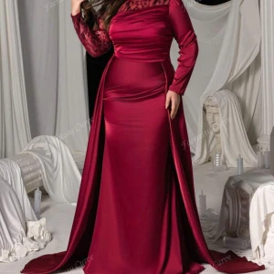 Vinröd långklänning med spetsdetaljer - Snygg vinröd långklänning med figurnära passform och långa ärmar. Överdelen har transparent spets med blommönster och axelpartiet är elegant. Kjolen är slät och har ett dramatiskt släp. Perfekt för bal eller fest, tillverkad i glansigt satinliknande material.