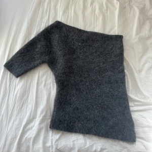 Grå stickad oneshoulder-tröja från Zara - Säljer en fluffig grå stickad tröja från Zara i oneshoulder-modell. Tröjan har en lång ärm och en axelbandslös sida, vilket ger en cool asymmetrisk look. Perfekt för dig som gillar att sticka ut med din stil och vill ha något mjukt och mysigt. Liten i storleken och endast använd en gång