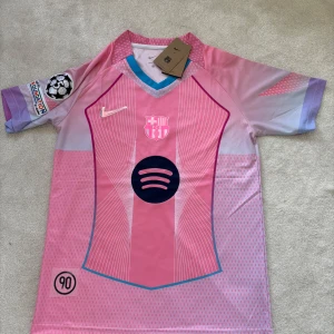 Rosa FC Barcelona Nike fotbollströja M - Snygg FC Barcelona fotbollströja från Nike i rosa och lila med korta ärmar. Tröjan har klubbmärke på bröstet, Spotify-logga, UEFA Foundation-patch och coolt grafiskt mönster. Tillverkad i lätt och ventilerande polyester, perfekt för match eller träning.