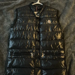 Svart dunväst från Moncler - Snygg svart dunväst från Moncler med glansig finish och klassisk logga på bröstet. Västen har hög krage, två dragkedjefickor framtill och quiltad design. Perfekt att slänga över en hoodie för en cool look.