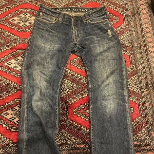 Blå jeans från American Eagle - Snygga blå jeans från American Eagle med klassisk femficksdesign och slitna detaljer framtill. Jeansen har normal passform och raka ben, perfekt för en avslappnad stil. Materialet är mjukt denim med tydliga slitningar för en cool look.