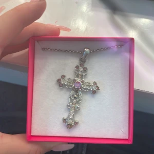 Korshalsband med rosa stenar SNÖ - Snyggt halsband från SNÖ of Sweden med ett stort kors som hänge, dekorerat med glittriga vita och rosa stenar samt en lila sten i mitten. Kedjan är silverfärgad med små kulor och ger en cool vibe. Perfekt för dig som gillar statement-smycken och detaljer.