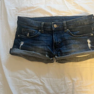 Mörkblå jeansshorts med slitningar - Snygga mörkblå jeansshorts med upprullade ben och slitna detaljer. Klassisk femficksmodell med knapp och dragkedja framtill. Passformen är tight och midjan är låg. Perfekta för varma dagar och ger en cool vibe till din outfit. Pris går att diskutera. Väldigt lågmidjade