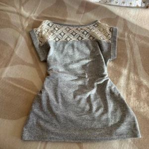 Grå T-shirt med spetsdetalj - Säljer en grå T-shirt med snygg vit spets upptill. Har korta ärmar och en rak, enkel passform. Spetsen har ett geometriskt mönster som ger en fin kontrast mot det mjuka grå tyget. Perfekt för dig som gillar stilrena detaljer.