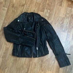 Svart skinnjacka från Lindex, strl 34 - Snygg svart skinnjacka från Lindex i storlek 34. Jackan har coola quiltade detaljer på ärmarna, flera dragkedjor och spännen i midjan. Använd fåtal gånger, säljes pga för liten