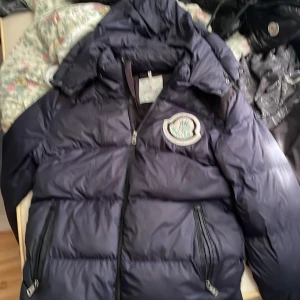 Mörkblå Moncler pufferjacka med huva - Säljer en mörkblå pufferjacka från Moncler med stor broderad logga på bröstet och huva. Jackan har två dragkedjeförsedda fickor och coolt serietidningsfoder på insidan. Perfekt för dig som vill ha en varm och snygg jacka med streetkänsla.köpte den på plick för 1000kr så värkar inte vara äkta men vet inget om den den är aldrig andvänd för den e för stor inga skador förutom lite innan för jackan vid monclere flisen som man kan se på bilden