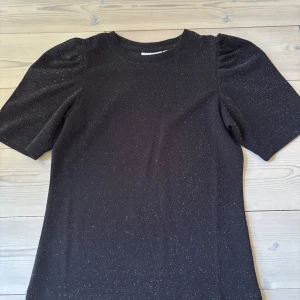 Svart glittrig topp från Kappahl XS - Svart topp från Kappahl i storlek XS med glittriga detaljer. Modellen har puffade korta ärmar och rund halsringning. Den är stretchig och passar till nästan allt.